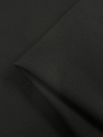 Close-up of a black **Super Cheap Fabrics Premium Viscose Suiting - Raven - 130cm** with a visible fold, showing the texture and smoothness of the material. The lighting highlights the fabric's matte finish and the precise seam where it is folded, ideal for suits and jackets.