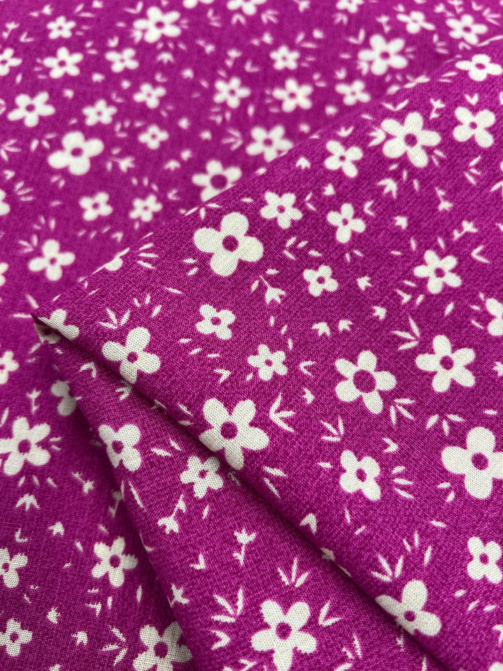 A close-up image of a beginner-friendly fabric with a purple background and a pattern of small white flowers. The lightweight fabric is folded neatly, creating layers that showcase the floral design. The texture appears soft with a slight sheen, ideal for **Super Cheap Fabrics** Bamboo Rayon - Pink DaisyRain - 145cm enthusiasts.