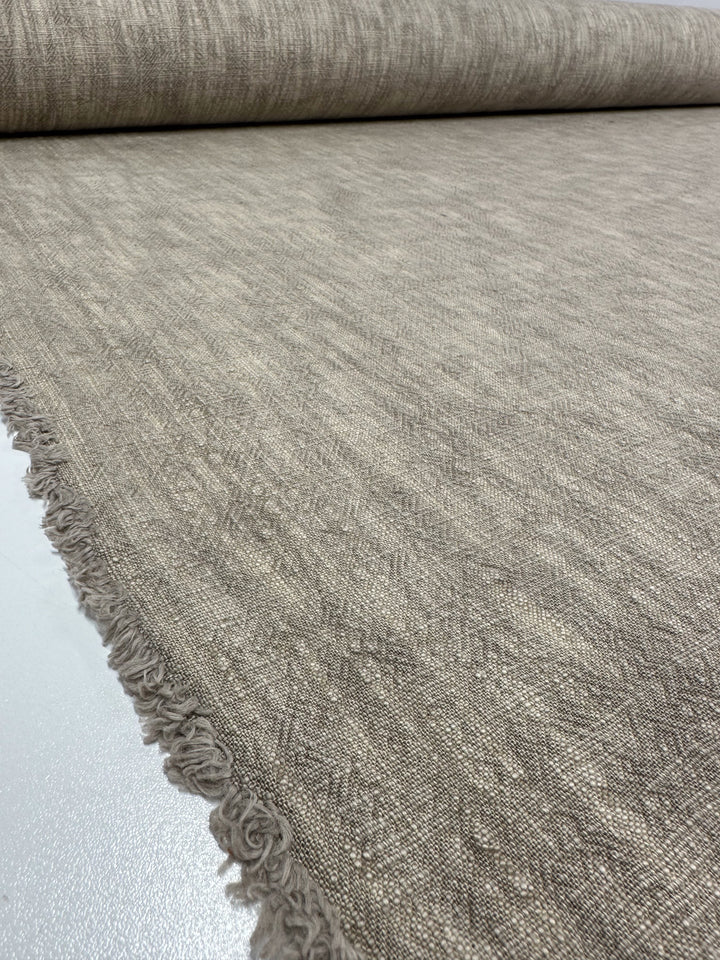 A close-up view of a roll of Basket Dyed Cotton - Taupe - 140cm from Super Cheap Fabrics, unfurled on a white surface. This light brown upholstery fabric features fringed edges and a subtle twill weave pattern, giving it a rustic appearance.