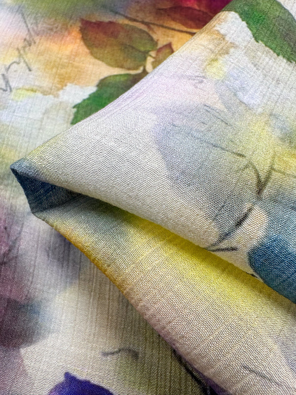 A close-up of a folded piece of the "Designer Cotton - Isabelle - 150cm" by Super Cheap Fabrics showcases a luxurious watercolor-style floral pattern. This high-quality fabric features soft hues of green, yellow, purple, and blue, along with visible texture and intricate details, making it perfect for luxury sewing projects.