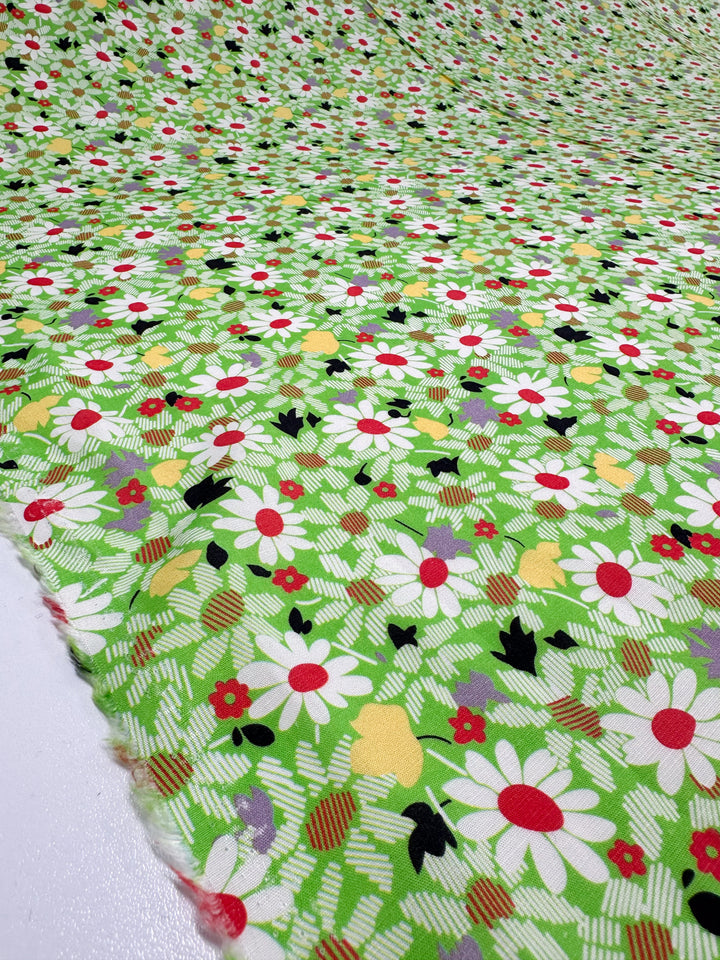 A close-up of Super Cheap Fabrics' "Printed Linen - Daised - 138cm" reveals a lively design featuring white daisies, red and yellow flowers, and black leaves on a green background. Ideal for garments or brightening up home decor.