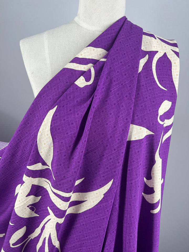 A mannequin is draped with the lightweight, purple Bamboo Rayon - Lily Shadows fabric from Super Cheap Fabrics. This 152cm fabric features a bold white floral design and is wrapped around the shoulder, showcasing its pattern and texture against a plain gray background, making it beginner-friendly for any new project.