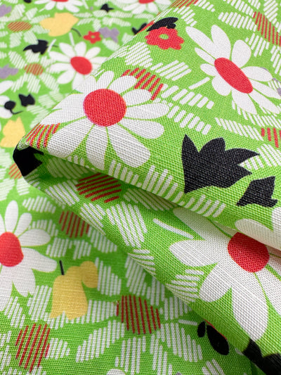 The close-up of the "Printed Linen - Daised - 138cm" by Super Cheap Fabrics displays a vibrant pattern with large white daisies, red centers, black leaves, and small yellow shapes on a bright green checkerboard background—ideal for garment creations or home decor.