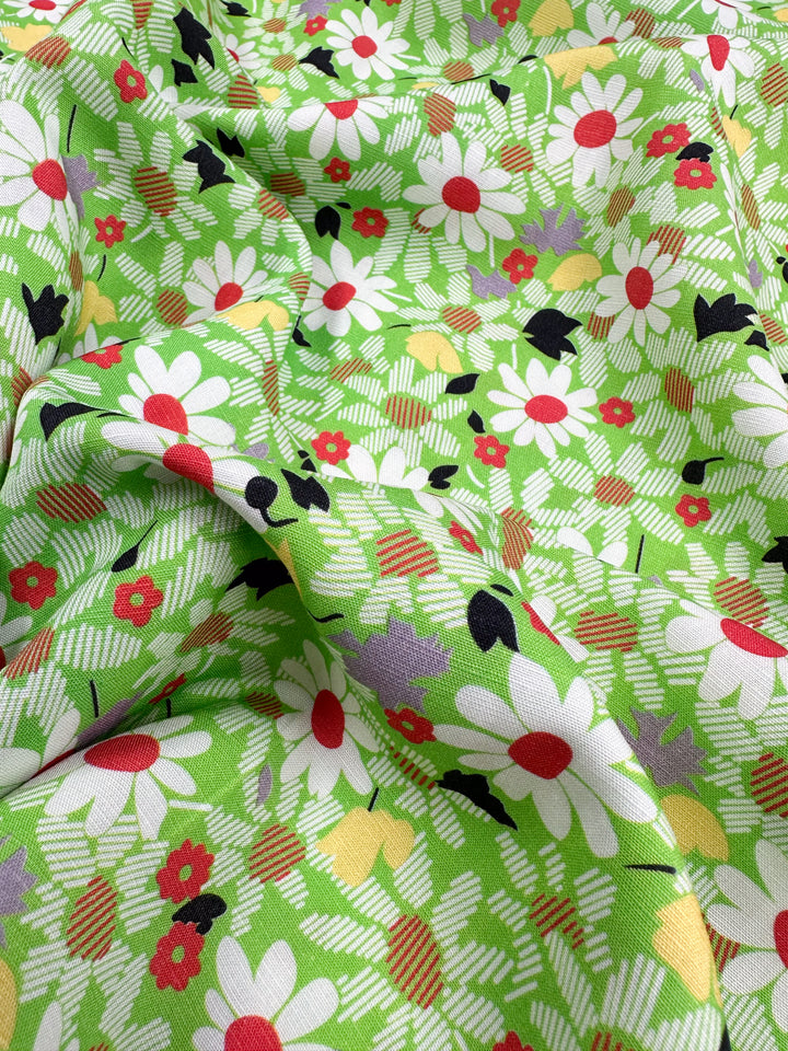 The Printed Linen - Daised by Super Cheap Fabrics features a vibrant floral pattern with large white daisies with red centers and small red and yellow flowers on a bright green background, accented with abstract black, white, and purple shapes. Perfect for garments or home decor. .