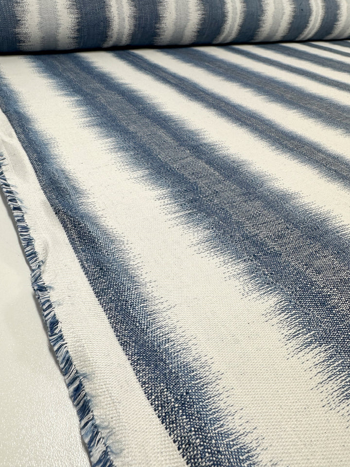 A close-up of the Upholstery Jacquard - Pacific by Super Cheap Fabrics highlights a gradient pattern of blue and white stripes. The soft yet durable texture features slightly frayed edges. Perfect for bringing elegance into your interiors. Width: 140cm.