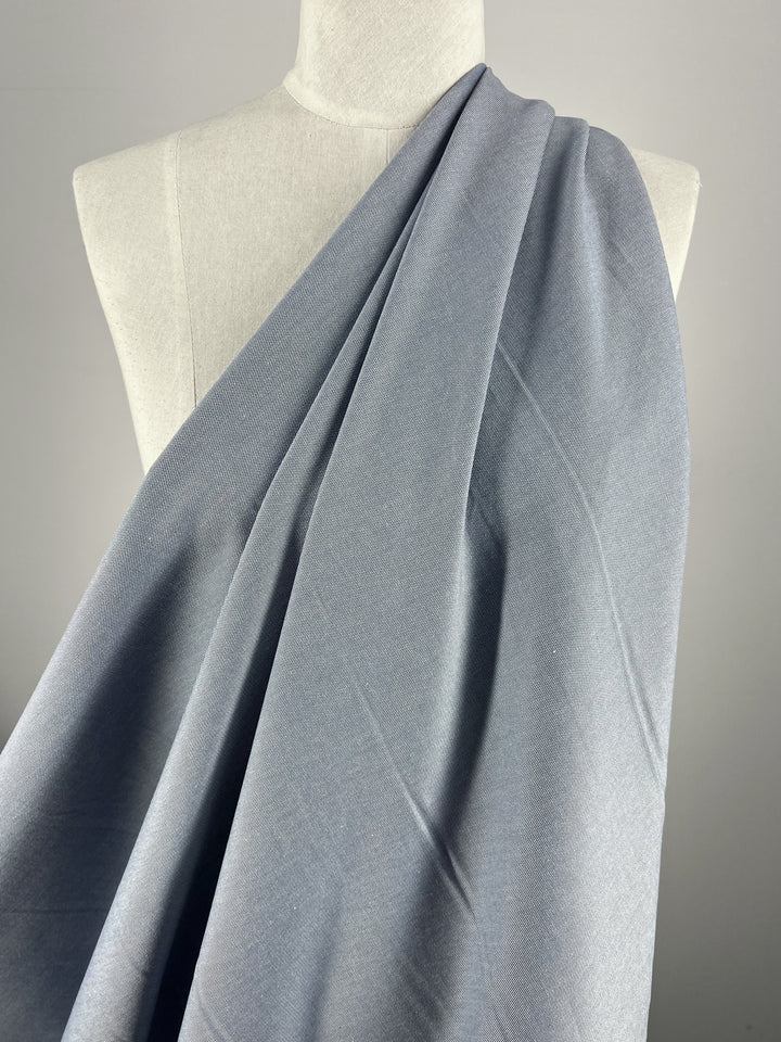 The Suiting - Eventide - 135cm from Super Cheap Fabrics elegantly drapes over a mannequin, highlighting its light gray hue with Silver Lake Blue hints. Its soft texture and smooth flow are perfectly showcased against a neutral backdrop, emphasizing its versatility.