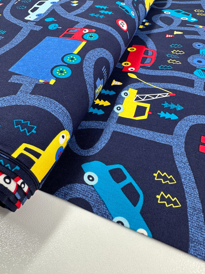 The Printed Cotton - Traffic Jams by Super Cheap Fabrics features cartoon vehicles like cars, trucks, and construction equipment on winding roads. A dark blue background highlights the vibrant red, yellow, and blue vehicles on this 112cm natural cotton fabric ideal for multi-use textiles.