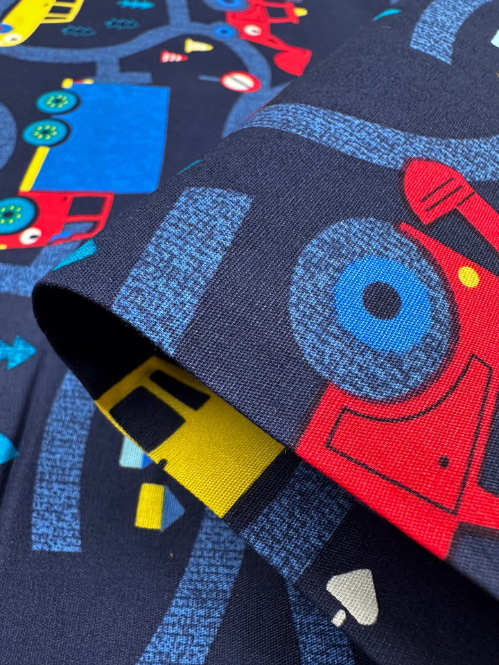 Close-up of Super Cheap Fabrics' Printed Cotton - Traffic Jams, 112cm, showcasing a vibrant pattern with blue and red trucks, yellow machinery, and road signs on a navy background for playful multi-use textiles.