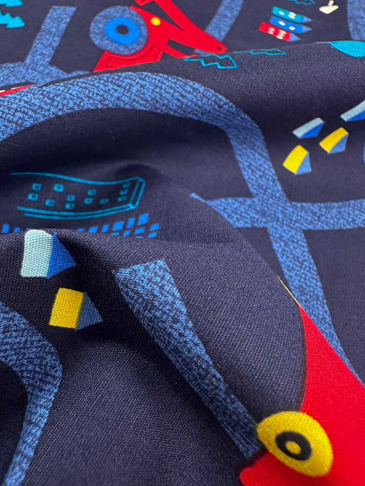 Close-up of Printed Cotton - Traffic Jams fabric by Super Cheap Fabrics displaying abstract geometric patterns in red, light blue, yellow, and teal on dark blue cotton. Slightly folded fabric creates a textured and layered look, perfect for multi-use textiles. Width: 112cm.