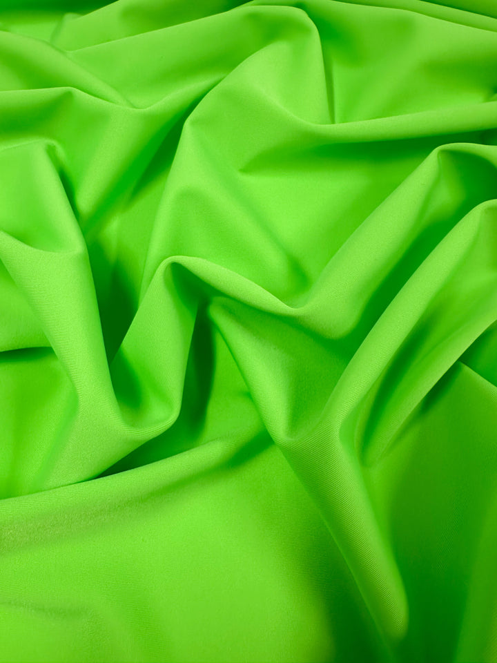 A close-up of the Nylon Lycra - Green Flash - 160cm fabric from Super Cheap Fabrics showcases its vibrant green color, with smooth folds and drapery creating an abstract pattern. The material looks soft and silky, with a four-way stretch that enhances the highlights and shadows, accentuating its luxurious texture.