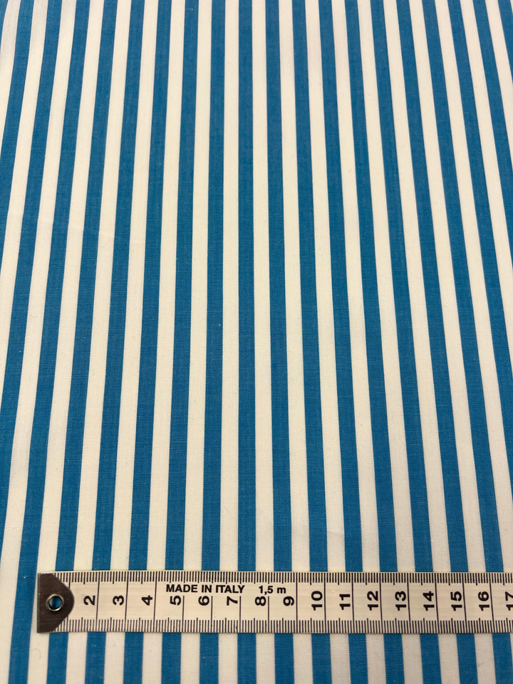 A lightweight cotton fabric with vertical white and turquoise stripes, known as Cotton Lawn - Vivid Blue - 150cm from Super Cheap Fabrics. A measuring tape at the bottom shows centimeter markings and reads "MADE IN ITALY," highlighting the fabric's pattern alignment, perfect for warmer weather.