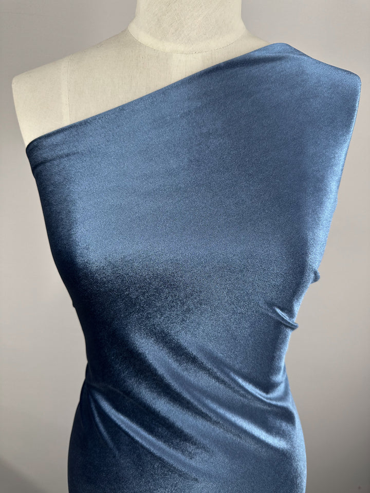 A blue velvet dress on a mannequin, crafted from Super Cheap Fabrics' Stretch Velour - Naval Academy. The one-shoulder design highlights the medium weight stretch fabric's smooth, shiny texture that drapes smoothly, with a plain background enhancing its exquisite color and form.