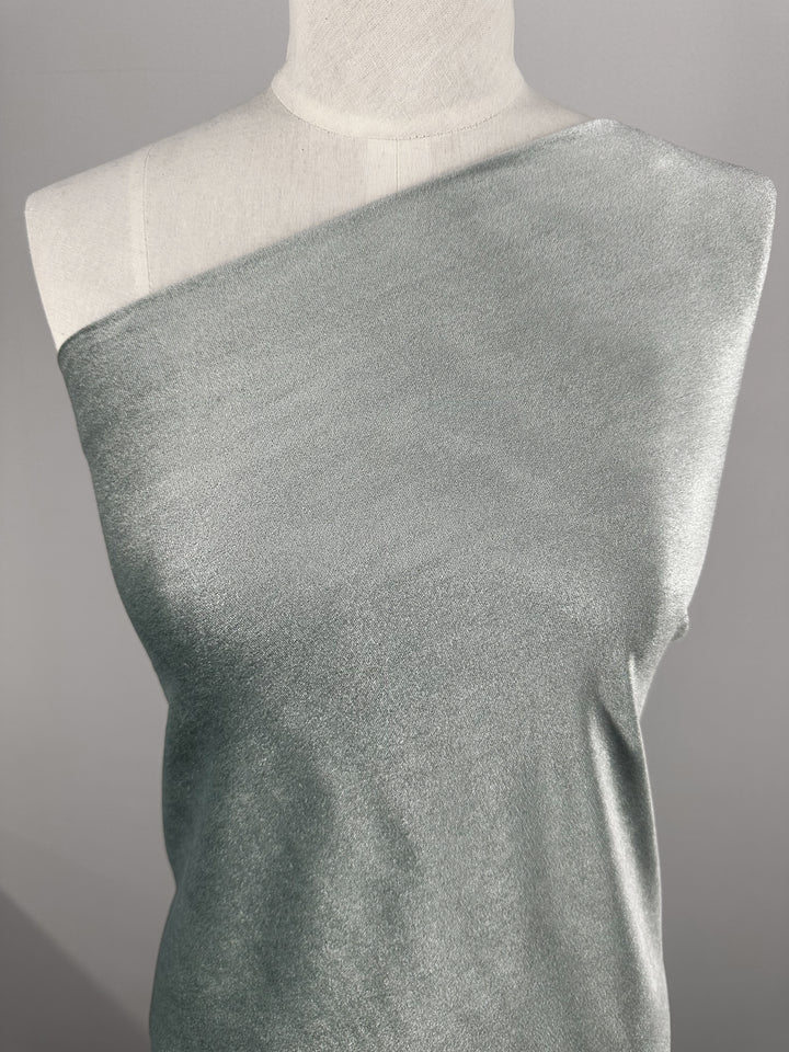 A mannequin is draped in Super Cheap Fabrics' Stretch Velour - High Rise - 170cm. This shimmering silver one-shoulder medium weight fabric boasts a smooth texture, subtly catching light. The plain gray backdrop enhances this modern stretch material's elegance.