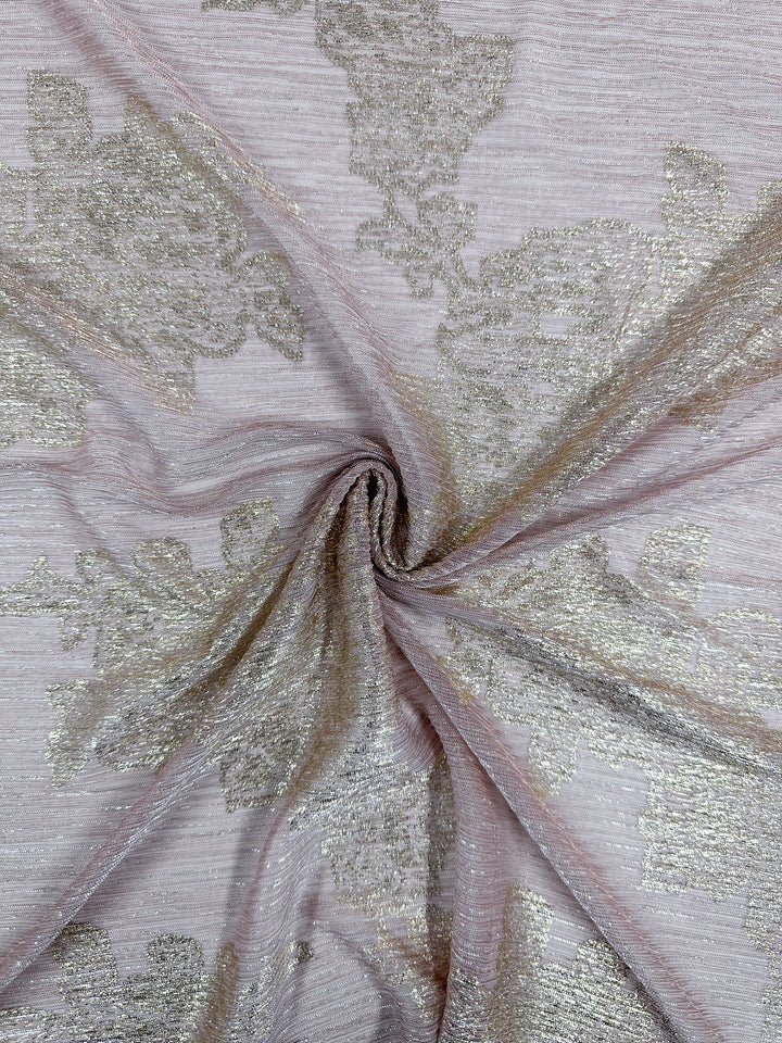 A close-up of the Metallix Pleat - Gold Leaf - 140cm fabric by Super Cheap Fabrics showcases its delicate baby pink hue with a swirling center. This lightweight polyester features an intricate, shimmering gold floral pattern. The fabric's texture appears soft and slightly translucent, highlighting its elegant design.