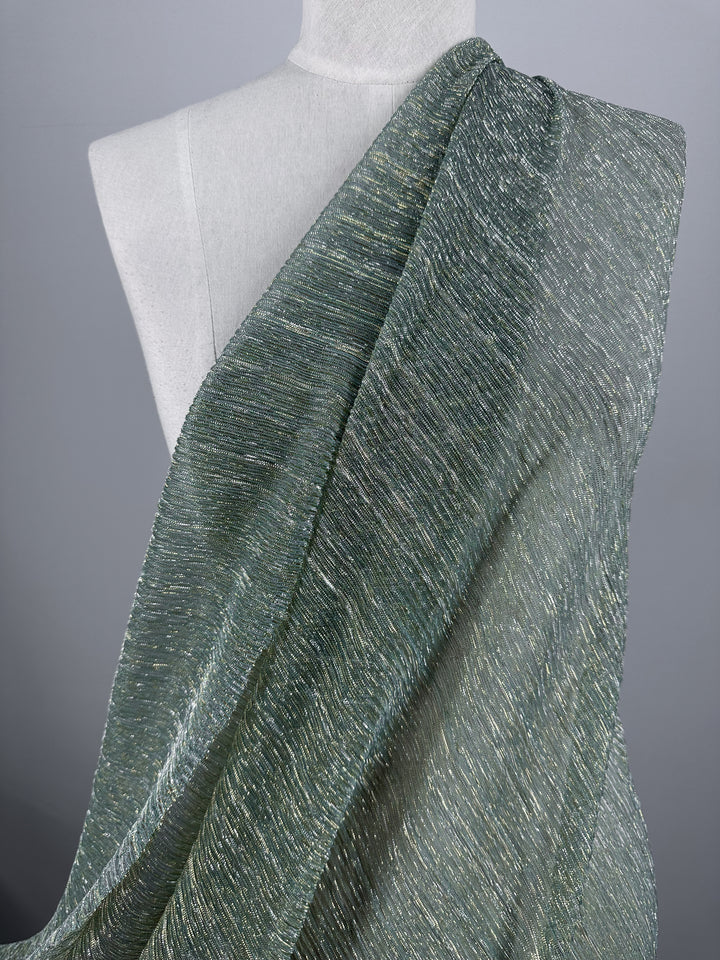 A mannequin is adorned with Metallix Pleat - Mermaid Glitter fabric from Super Cheap Fabrics. This shimmery green polyester material elegantly wraps around the shoulder and drapes down, showcasing its intricate woven pattern. The plain grey background enhances the fabric's lustrous sheen.