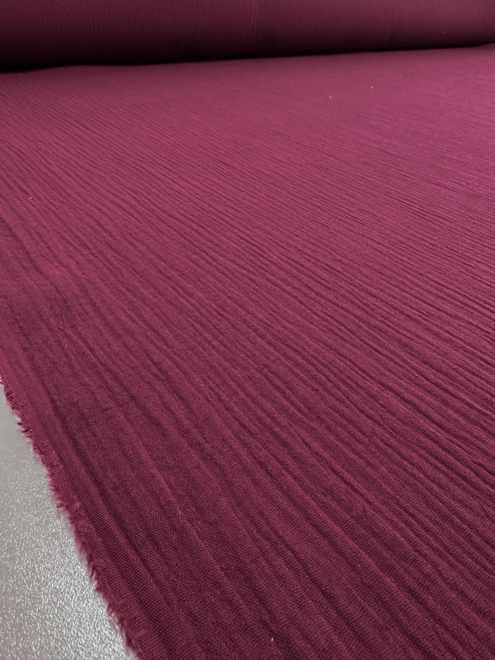 A large roll of Super Cheap Fabrics' Muslin in Rumba Red, measuring 135cm wide, lies flat on a surface, showcasing its grainy pattern and vibrant hue. The lightweight fabric stretches out with slightly frayed edges, emphasizing its natural texture.