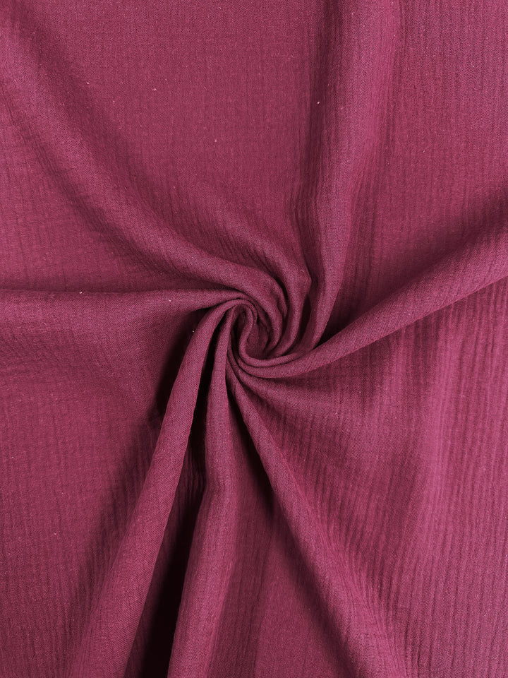 A close-up of crinkled Muslin - Rumba Red - 135cm from Super Cheap Fabrics reveals a swirling pattern at the center, creating a dynamic texture and spiral design. This lightweight, organic fabric appears soft and textured, with subtle lighting highlighting its folds.