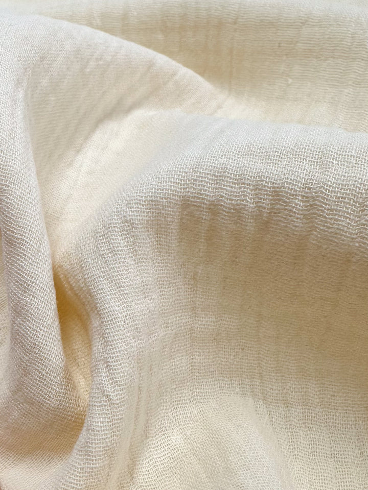 The sentence can be rewritten as follows:

A close-up of Super Cheap Fabrics' Muslin in Almond Milk, measuring 135cm, reveals its cream-colored, textured finish with a slightly crumpled appearance. The visible weave pattern showcases the fabric's softness and delicate texture, creating gentle folds and shadows.