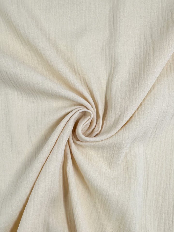 Close-up of the Muslin - Almond Milk - 135cm by Super Cheap Fabrics, showcasing its light beige organic fabric with a twist pattern at the center. The texture is soft, featuring gentle folds that radiate from the middle to create a subtle spiral effect. This versatile muslin is ideal for reusable applications, offering both elegance and style.