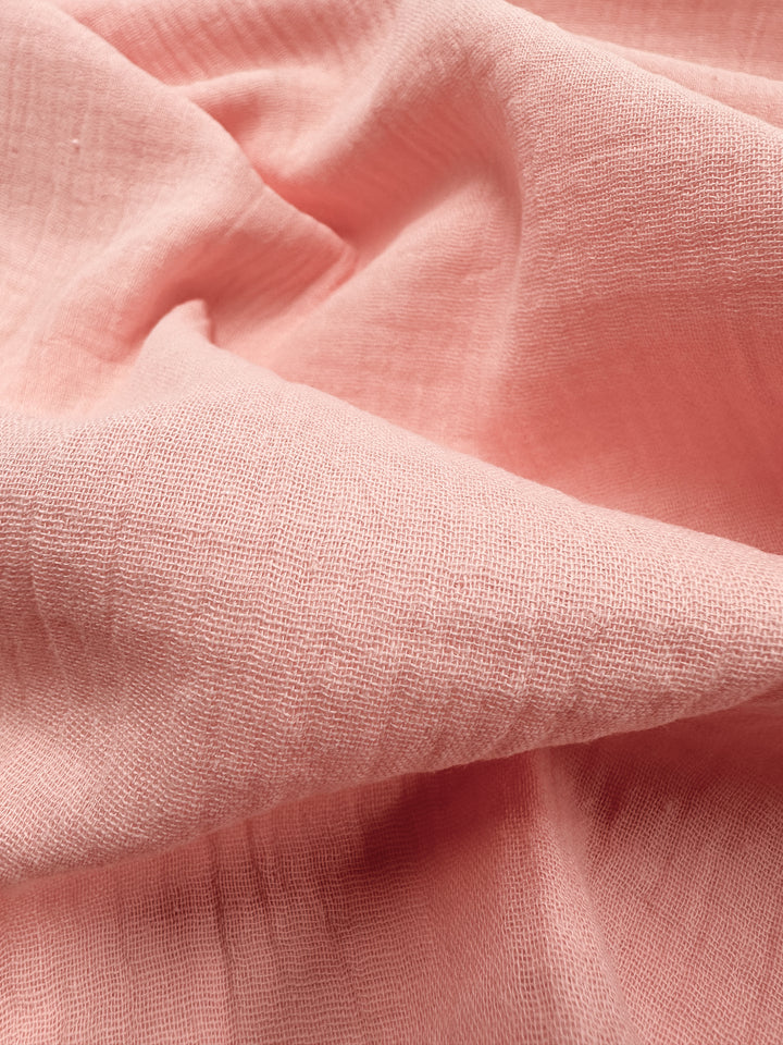 Close-up of soft, textured muslin fabric in the Peach Parfait color from Super Cheap Fabrics, featuring gentle folds and creases. The organic material is lightweight and slightly translucent at 135cm wide, capturing the light to highlight its smooth surface and subtle woven pattern.