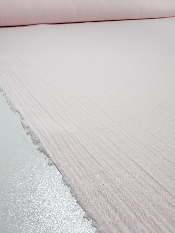 A close-up view of a roll of the "Muslin - Slightly Pink - 135cm" fabric by Super Cheap Fabrics, featuring its textured surface and frayed edge. The organic cotton fabric is displayed on a smooth, light-reflective surface.