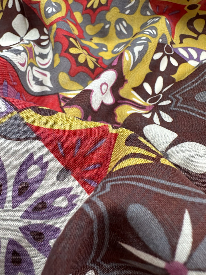 A close-up of the Linen Blend - Mosaic fabric by Super Cheap Fabrics reveals its lightweight texture adorned with a vibrant floral and geometric pattern. The design showcases abstract shapes in shades of red, yellow, purple, and brown, complete with swirling motifs. The breathable material has a soft, slightly crumpled appearance.