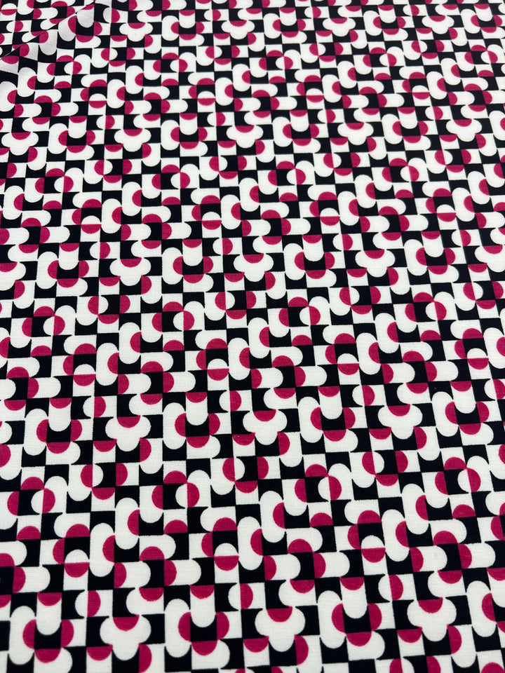 Super Cheap Fabrics' "Printed Lycra - Mirrors - 150cm" features interlocking black, white, and pink geometric shapes with semicircles and rectangles. This medium-weight fabric is visually striking and made from a comfortable polyester spandex blend.