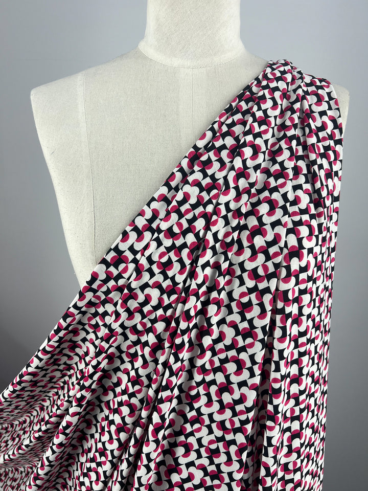 Draped on a mannequin, the Super Cheap Fabrics' Printed Lycra - Mirrors 150cm medium-weight fabric showcases a modern, eye-catching geometric pattern. Made of polyester spandex, it features bold red, black, and white curved shapes for a stylish look.