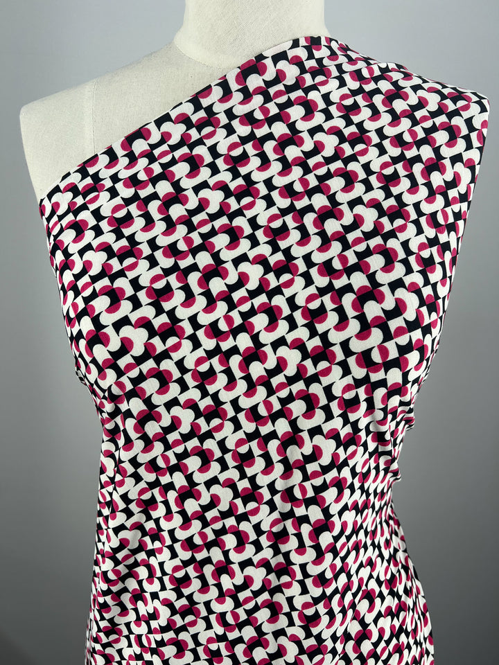 The dress mannequin showcases a "Mirrors" one-shoulder garment made from Super Cheap Fabrics' medium-weight Printed Lycra, featuring a geometric pattern with interlocking black, pink, and white shapes on a light background. Width: 150cm.