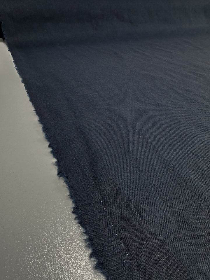 A close-up of Super Cheap Fabrics' Pure Washed Linen in India Ink (140cm) shows a textured dark blue roll on a light gray surface. Frayed edges are visible, and subtle shadows highlight the weave.