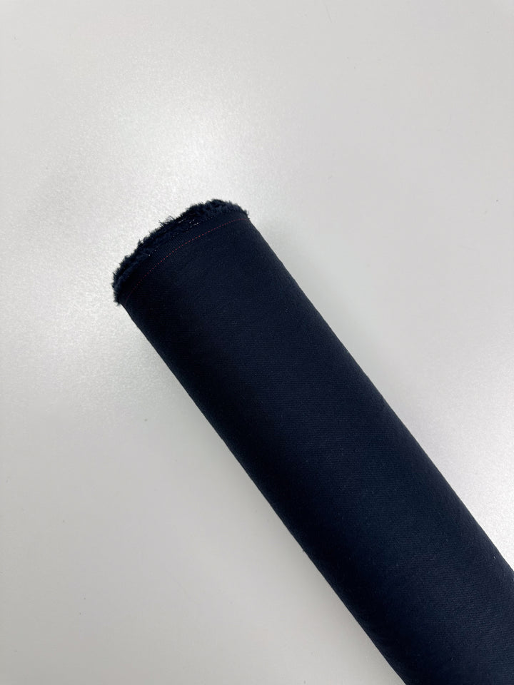 A roll of Super Cheap Fabrics' Pure Washed Linen in India Ink, 140cm wide, lies diagonally on a white surface. The smooth, tightly wound fabric has a frayed edge at one end, suggesting its suitability for crafting fine garments.