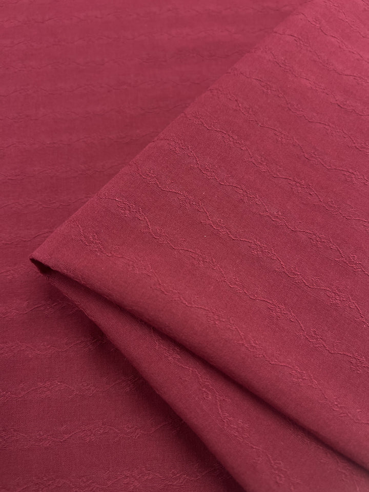 Close-up of a folded piece of Super Cheap Fabrics' "Textured Cotton - Red Plum - 145cm", showcasing its 100% cotton texture with a subtle wave pattern. The extra lightweight, soft fabric displays gentle shade variations due to lighting.