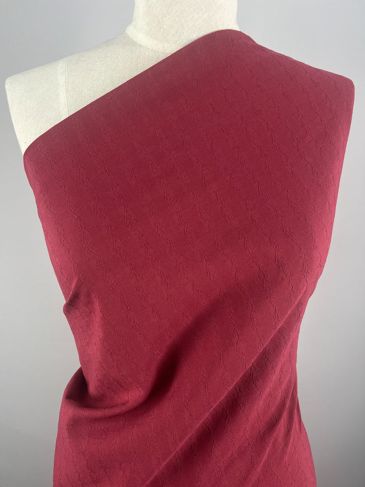 A mannequin displays Super Cheap Fabrics' Textured Cotton in vibrant Red Plum. The 100% cotton is draped one-shoulder against a gray background, featuring a subtle pattern and an extra lightweight feel. Width: 145cm.