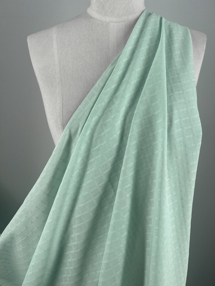 A draped piece of Textured Cotton - Honeydew from Super Cheap Fabrics is elegantly showcased on a neutral-colored dress form against a gray background. The light teal, extra lightweight cotton fabric features a subtle diamond pattern, and its texture and gentle folds highlight the 100% cotton fibers beautifully.