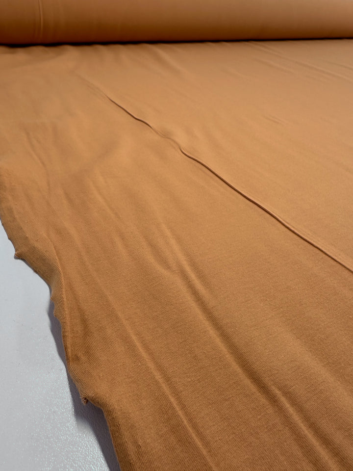 A close-up of Super Cheap Fabrics' Cotton Lycra in Caramel, 165cm wide, shows a medium weight, light brown fabric spread smoothly with minor wrinkles, ideal for bodycon dresses. It appears unrolled from a roll at the image's top edge.