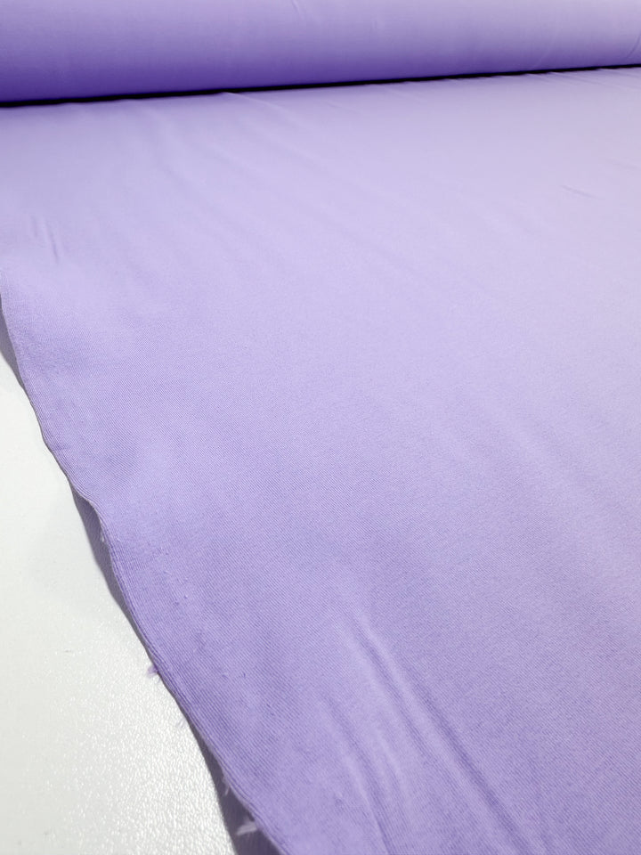 A large roll of Super Cheap Fabrics' Cotton Lycra - Purple Rose - 165cm is spread out on a flat surface, featuring its medium-heavy weight and breathable texture.