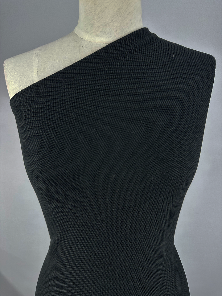 A beige mannequin torso is styled in a black, one-shoulder garment made from Super Cheap Fabrics' Waffle Knit - Black - 152cm fabric. The outfit envelops the entire chest and flows down the torso, highlighting its asymmetrical design with a delicate three-dimensional texture. The setting is a neutral gray background.