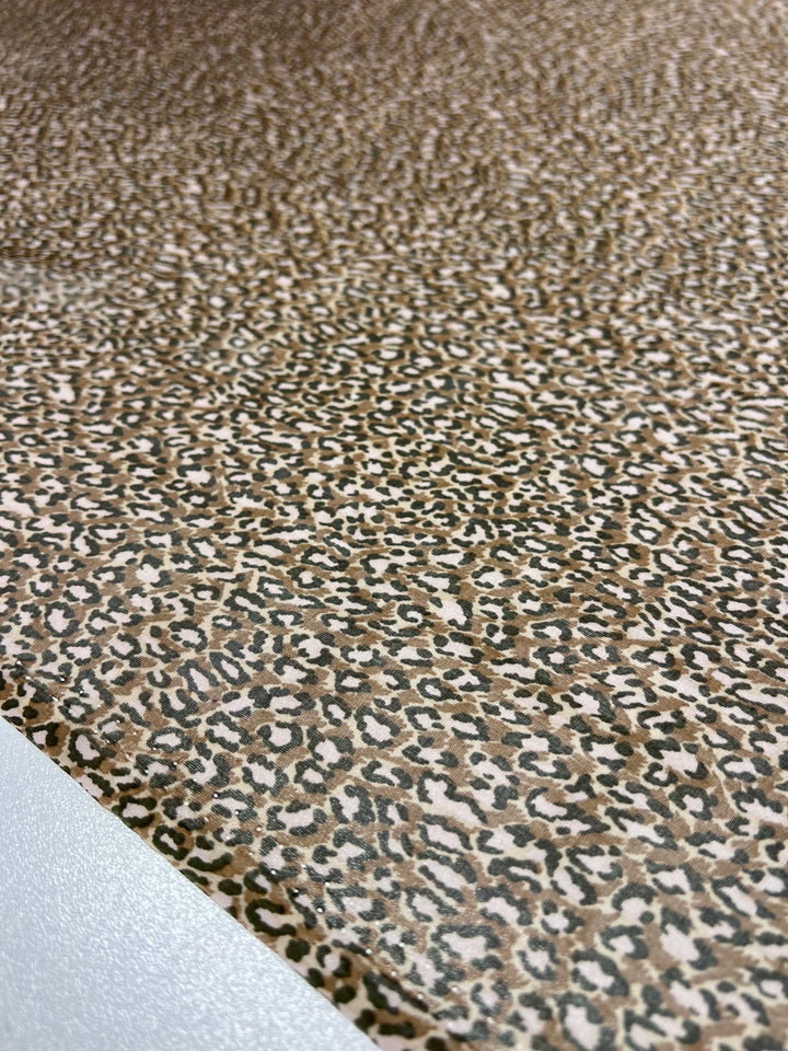 A detailed close-up of the Pure Printed Silk - Namir from Super Cheap Fabrics, showcasing a leopard print pattern with black and brown spots on a beige background. The soft 75 GSM fabric lies flat across most of the frame, revealing just a small section of a white surface in the bottom-left corner.