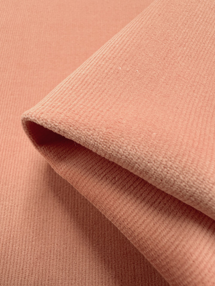 Close-up of Super Cheap Fabrics' Micro Wale Corduroy - Peach, 145cm wide, with visible vertical ridges showcasing its ribbed texture and soft appearance. Ideal for winter clothing, the fabric unfolds smoothly, highlighting its gentle color and pattern.