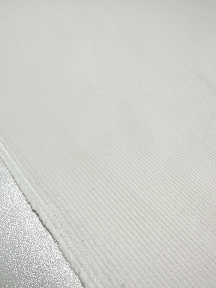 A close-up of Super Cheap Fabrics' Pin Wale Corduroy in Ivory reveals a textured white surface with subtle vertical ridges. The lighting creates gentle shadows that enhance the depth and dimension, akin to corduroy fabric. Width: 150cm.