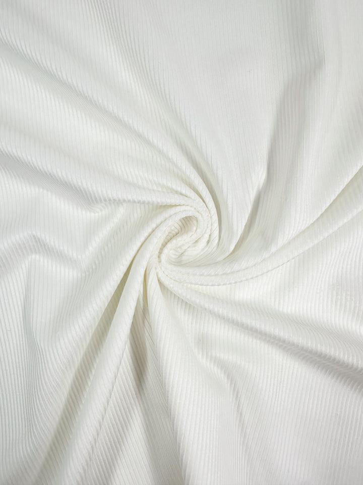 A close-up of Super Cheap Fabrics' Pin Wale Corduroy in Ivory shows its light, ribbed texture with fine vertical lines. The soft swirl pattern and clean, bright white color evoke cozy winter essentials.