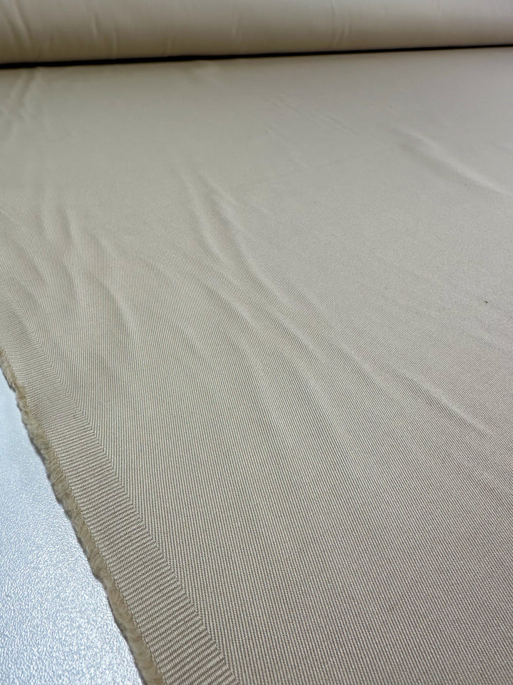 A close-up of a roll of Super Cheap Fabrics' Linen Drill - Ancient Scroll reveals its subtle creases, soft sheen, and finely woven heavy weight drill weave in light beige cotton spread on a surface.