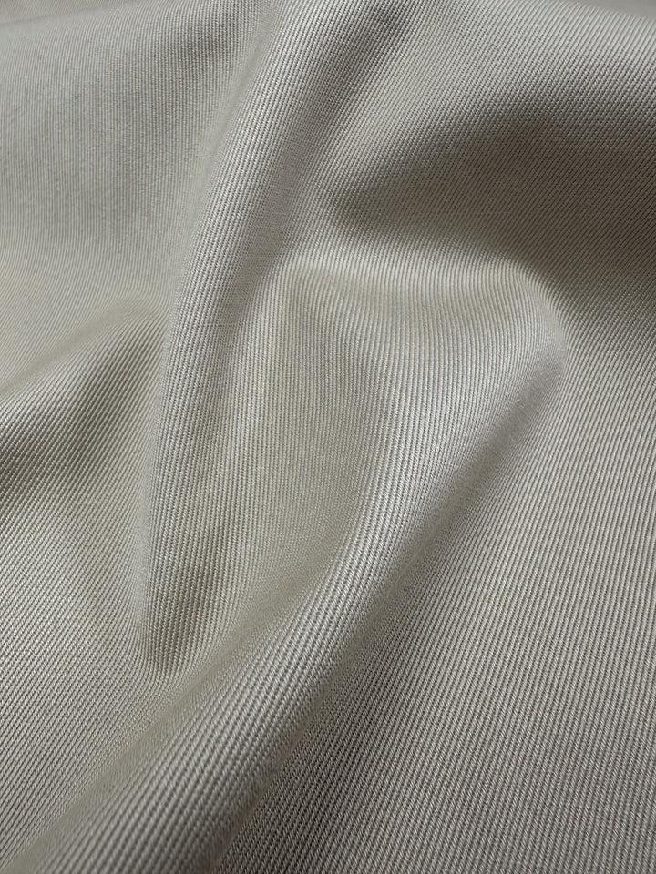 A close-up of Super Cheap Fabrics' Linen Drill - Ancient Scroll, 140cm, reveals its smooth light gray texture with a subtle diagonal ribbed pattern. Gentle folds in the fabric create soft shadows and highlights, enhancing its delicate weave and sheen.