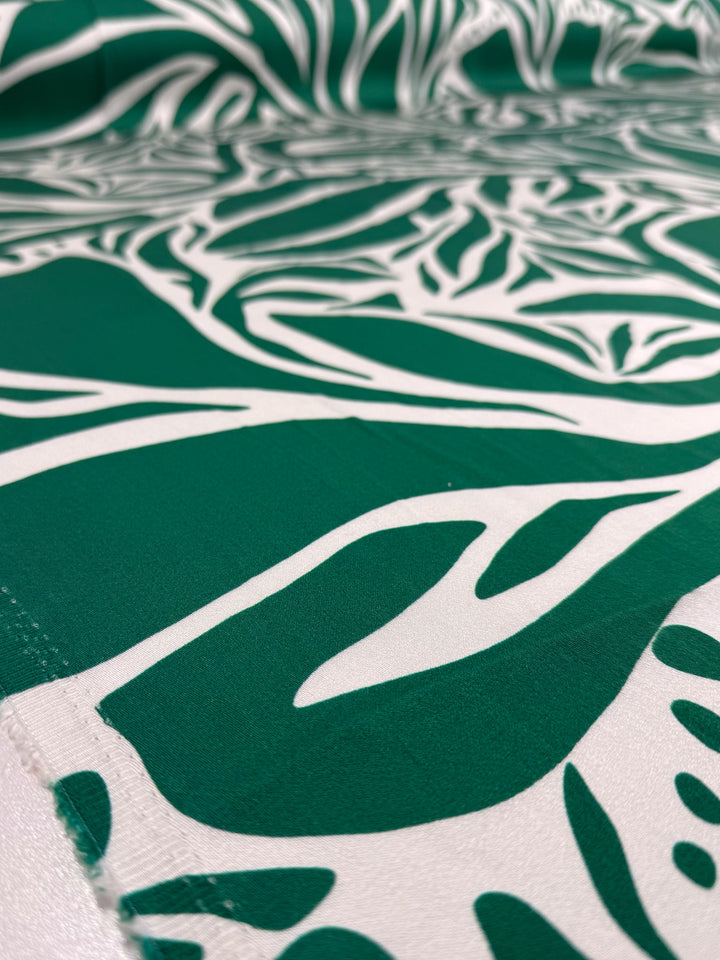 Close-up of Super Cheap Fabrics' Mercerised Rayon - Tropic Cutout (150cm), featuring a bold green and white abstract leafy pattern. The lightweight fabric is slightly folded, showcasing its smooth texture and detailed design.