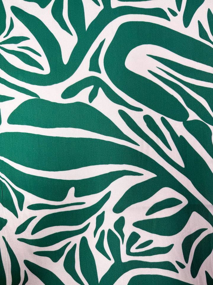 Mercerised Rayon - Tropic Cutout - 150cm by Super Cheap Fabrics features bold, dark green organic shapes on a white background, resembling stylized leaves. The lightweight fabric is smooth, breathable, and perfect for dynamic, nature-inspired garments.