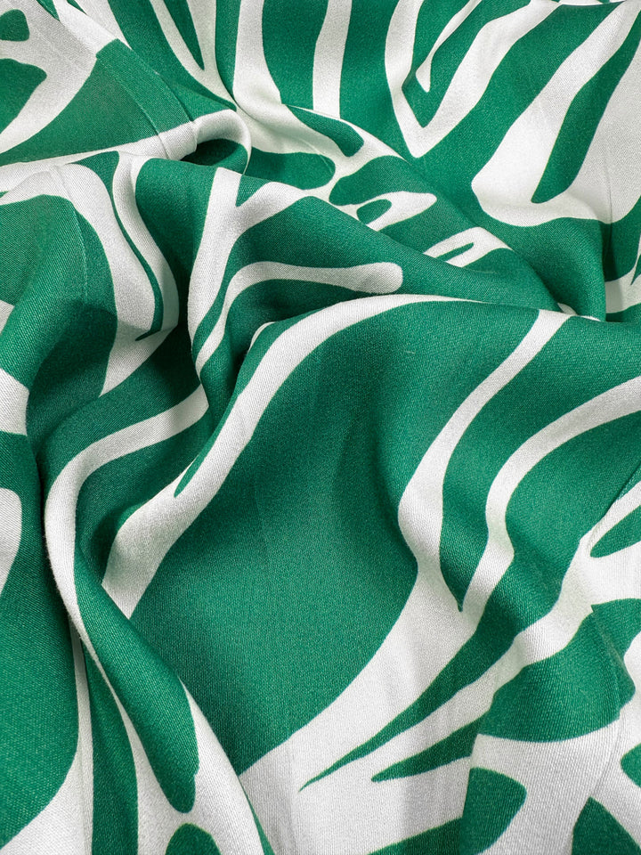 The Mercerised Rayon - Tropic Cutout by Super Cheap Fabrics features a bold green and white abstract leaf or zebra stripe pattern on breathable rayon, softly draping to form elegant folds and shadows. Width: 150cm.