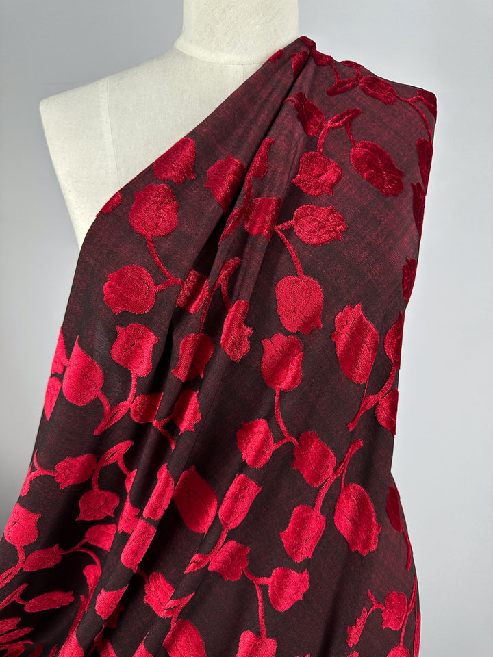 The mannequin is draped with Super Cheap Fabrics' luxurious Burnout Velvet - Lychee, 150cm, in rich burgundy, adorned with red embossed tulips. The fabric covers one shoulder, highlighting its intricate texture and vibrant color as a stunning piece of textile art.