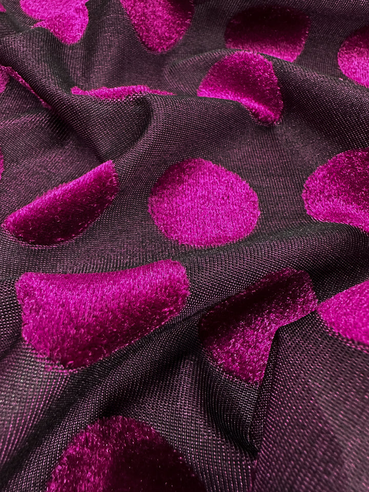 Close-up of Super Cheap Fabrics' Burnout Velvet - Festival Fuchsia, showcasing a luxurious fabric with a dark base featuring vibrant, fuzzy magenta polka dots evenly spread across its 155cm width, creating a bold and eye-catching pattern.