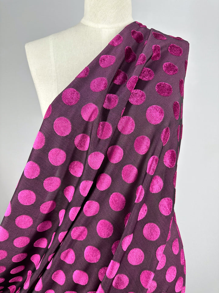 A mannequin displays the luxurious Burnout Velvet - Festival Fuchsia from Super Cheap Fabrics, featuring vibrant pink polka dots. The soft, velvety texture stands out against a plain gray background, highlighting its bold design.