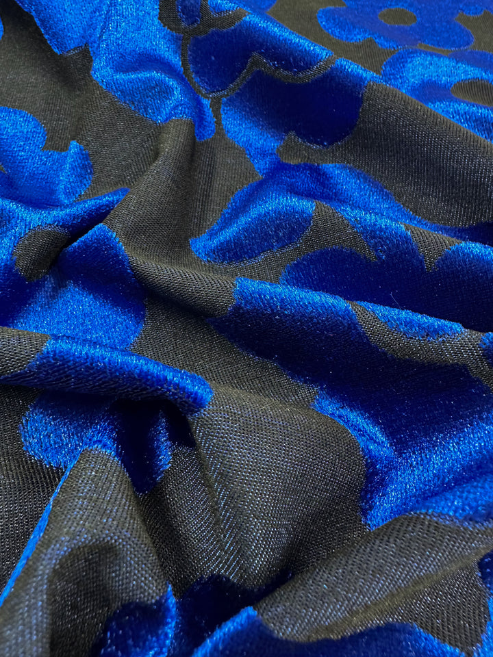 Close-up of Super Cheap Fabrics' Burnout Velvet - Snorkel Blue - 155cm, showcasing its luxurious fabric with a vivid blue floral pattern on black. The soft, slightly wrinkled texture highlights the vibrant textile design.